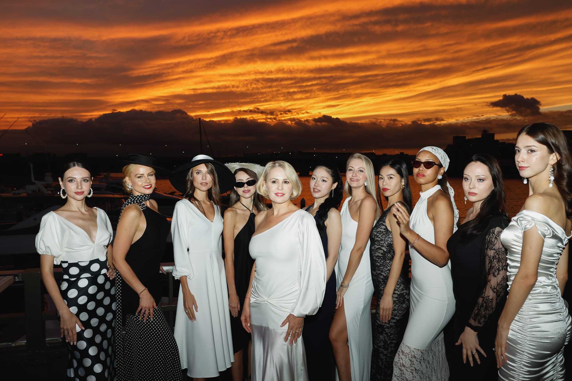 夕焼けのマリーナで、白と黒のドレスをまとった11人のモデルが並ぶ集合写真