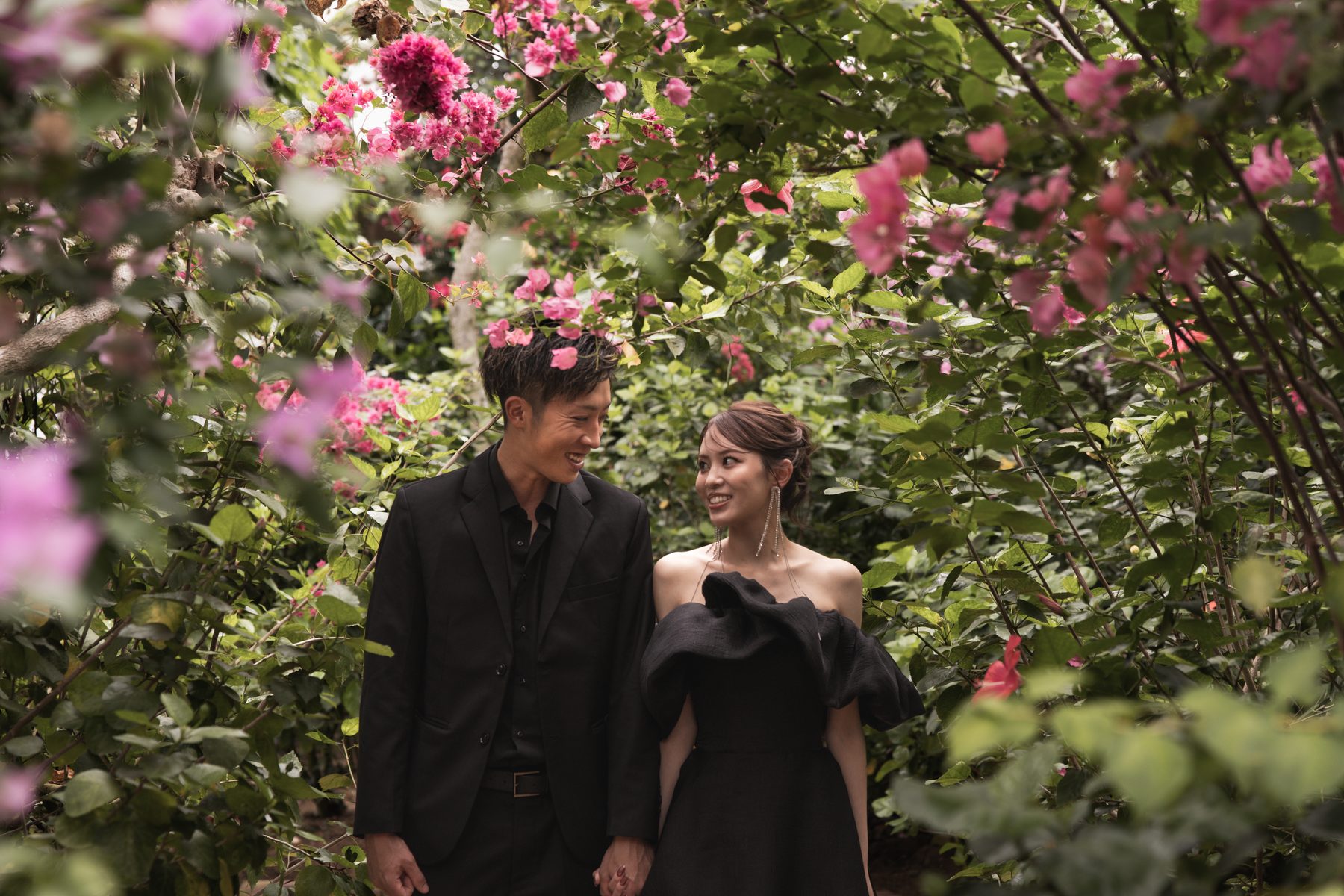 黒を着たカップルが、ピンクの花に囲まれた庭を歩きながら見つめ合う